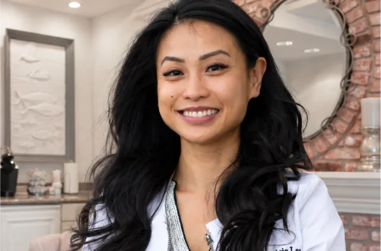 Dr. Patricia Lee with long dark wavy hair smiling warmly in a white lab coat embroidered with her name at Dental Expressions, with a brick wall and decorative mirror in the background